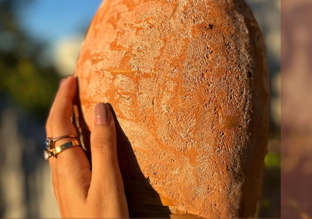 A hand holding a handmade earth-plastered piece, its textured orange surface lit by sunlight